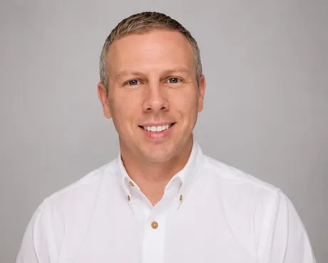 A man with short fair hair in a white button-down shirt smiles against a plain grey studio backdrop.