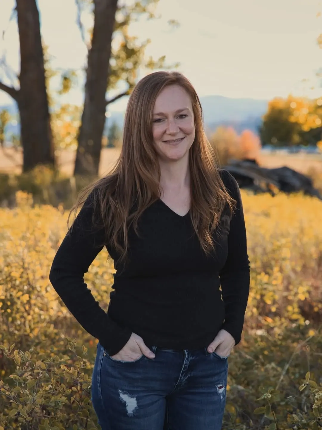 A woman with long auburn hair in a black V-neck sweater and jeans stands with her hands in her pockets in an autumn field.