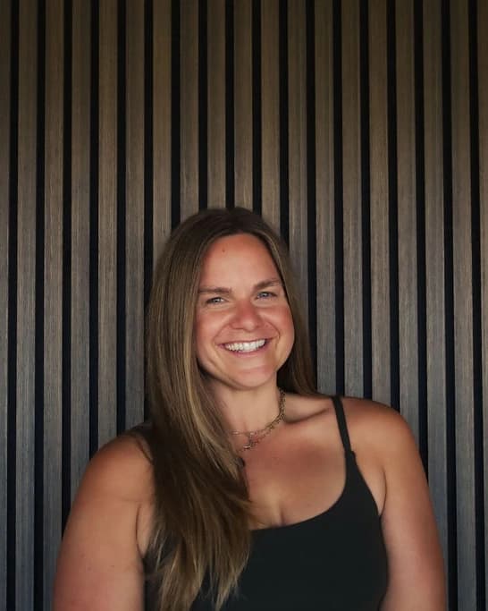 A woman with long straight light-brown hair, a black camisole and a fine chain necklace smiles in front of a slatted wood wall.