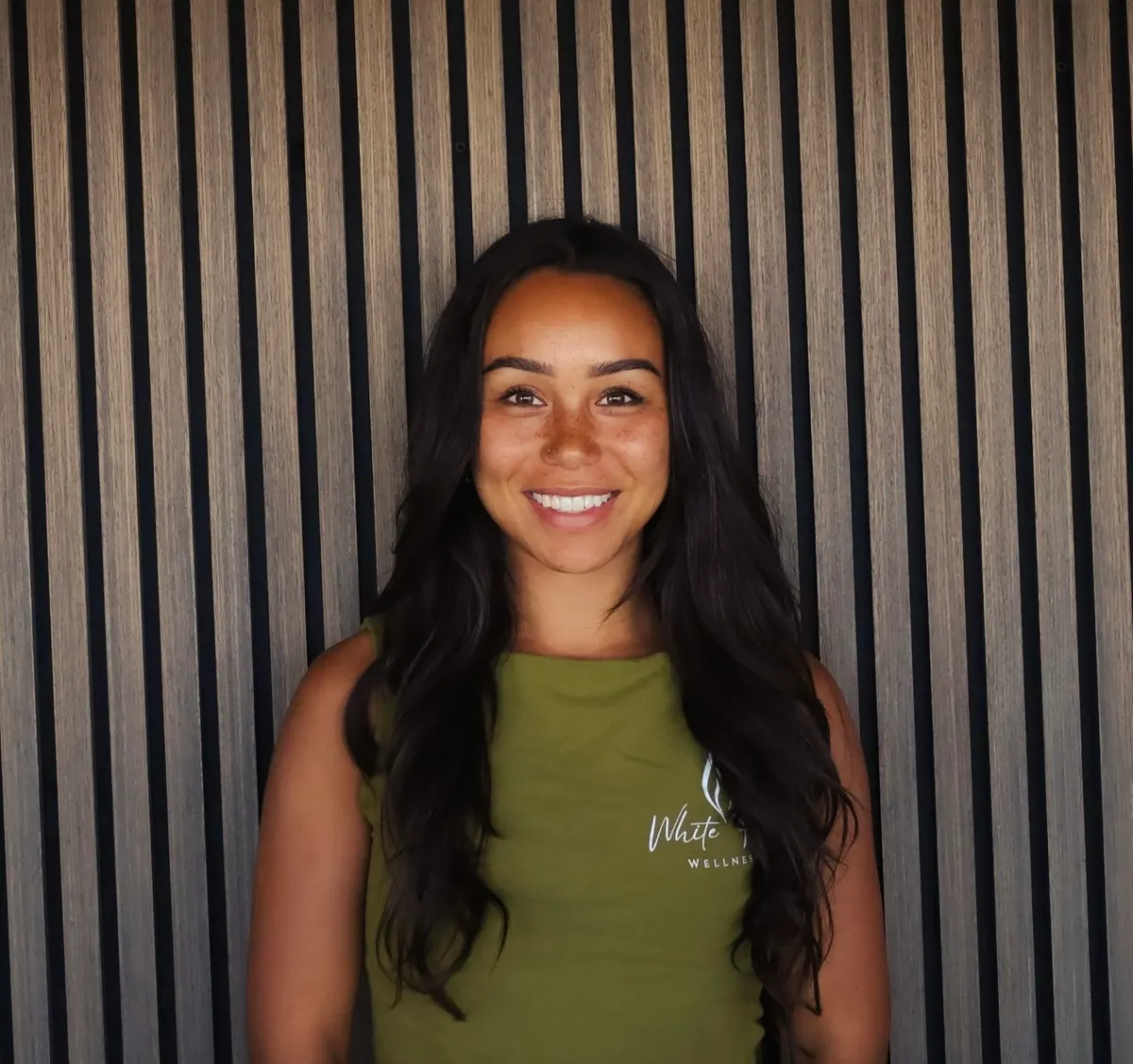 A woman with long dark wavy hair in an olive tank top smiles in front of a slatted wood wall.