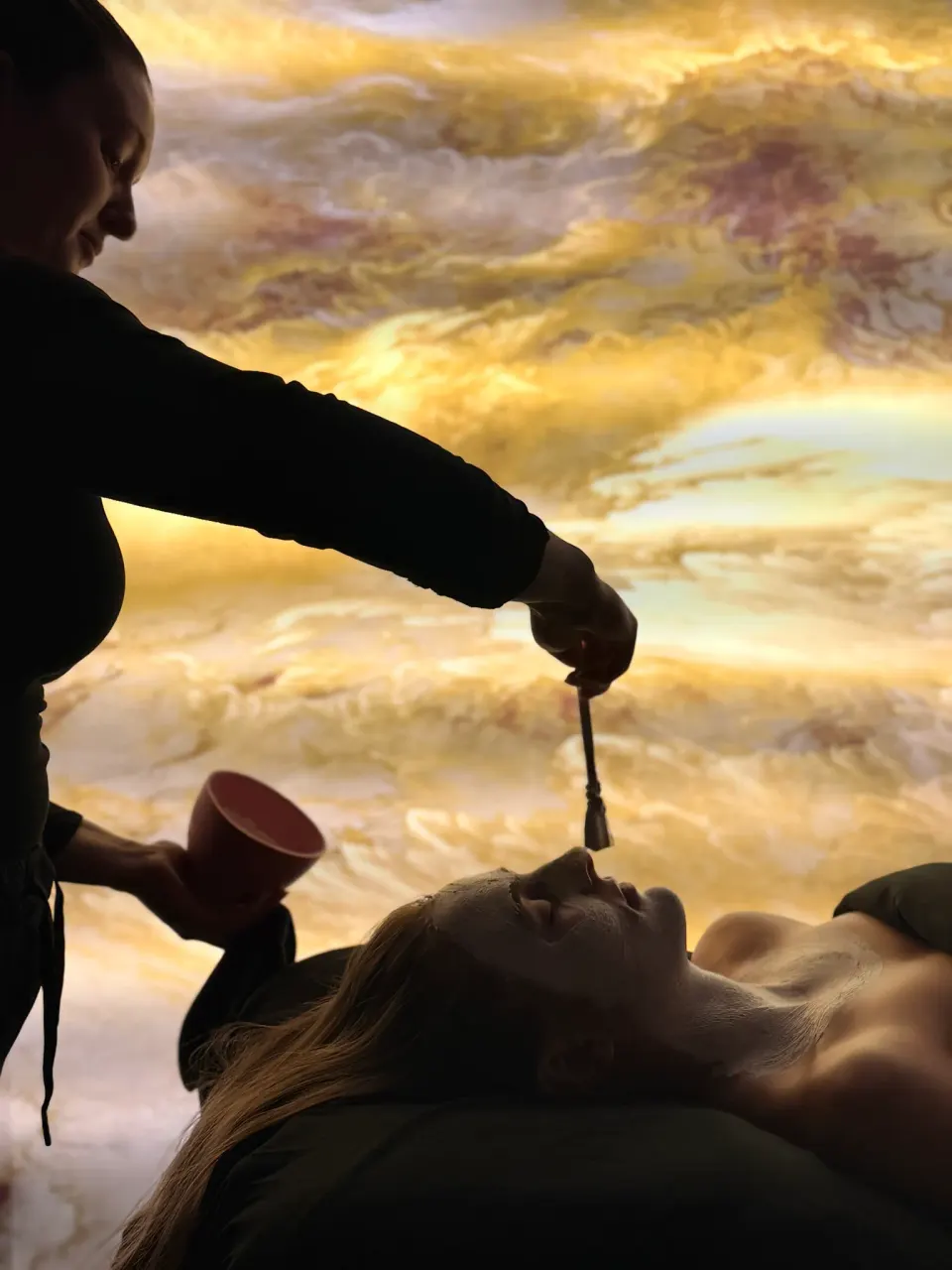 An esthetician brushes a clay mask onto the face of a client lying back, a bowl in her other hand, against a lit gold stone wall.