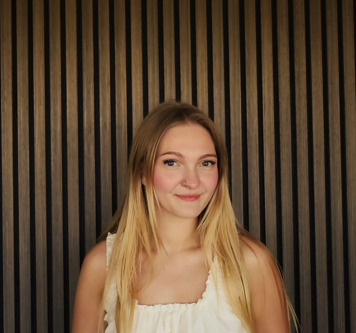 A woman with long blonde hair in a cream off-shoulder top smiles in front of a slatted wood wall.
