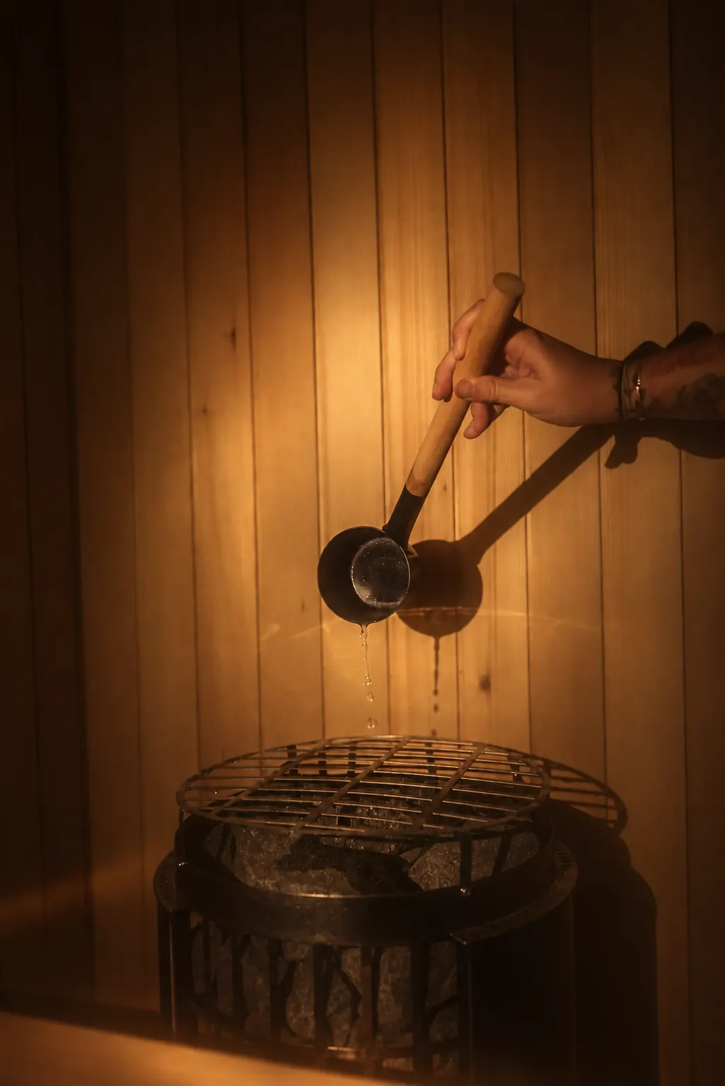 A hand tips a long-handled ladle over the stones of a sauna stove, water falling in a thin stream, against warm-lit hemlock boards.