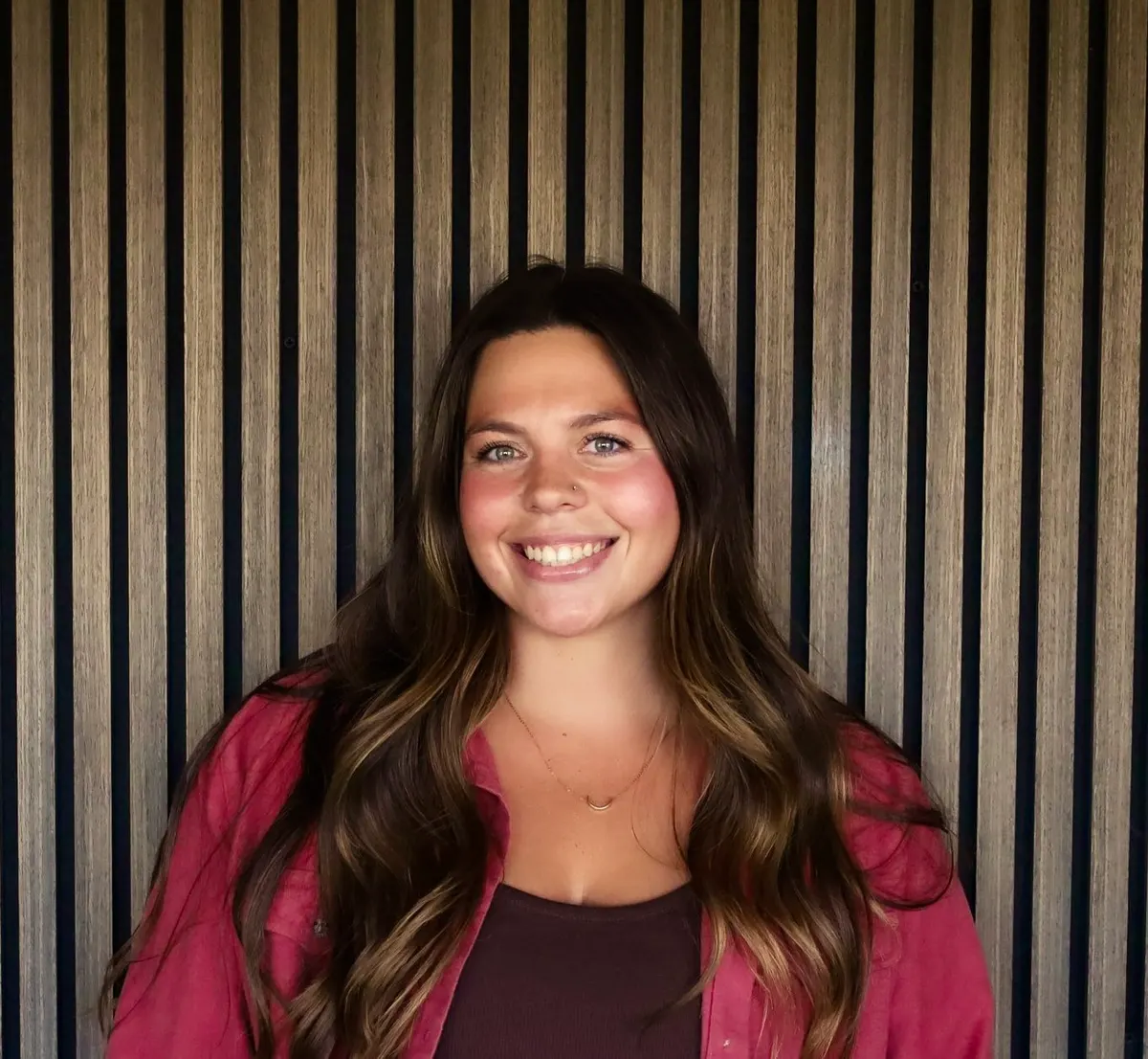 A woman with long brown hair in a raspberry jacket smiles in front of a slatted wood wall.
