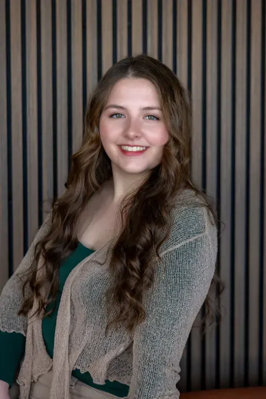 A woman with long wavy brown hair, a green top and a loose-knit tan cardigan smiles in front of a slatted wood wall.