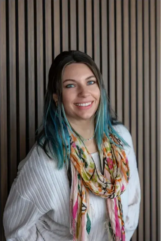 A woman with dark hair dyed teal toward the ends, a white ribbed sweater and a floral scarf smiles in front of a slatted wood wall.