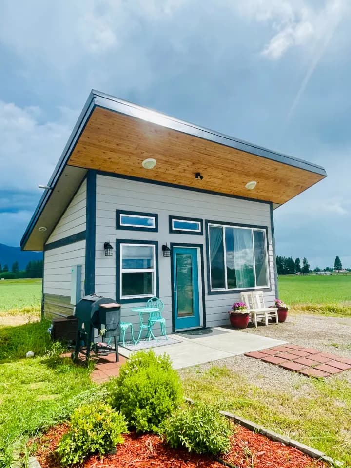 A small gray cabin with navy trim, a teal front door and a wood-lined roof overhang, in green fields under a stormy sky.