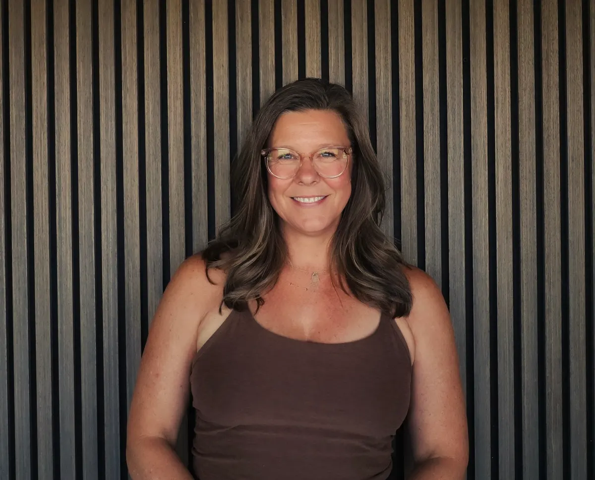 A woman with long greying hair and glasses in a brown tank top smiles in front of a slatted wood wall.