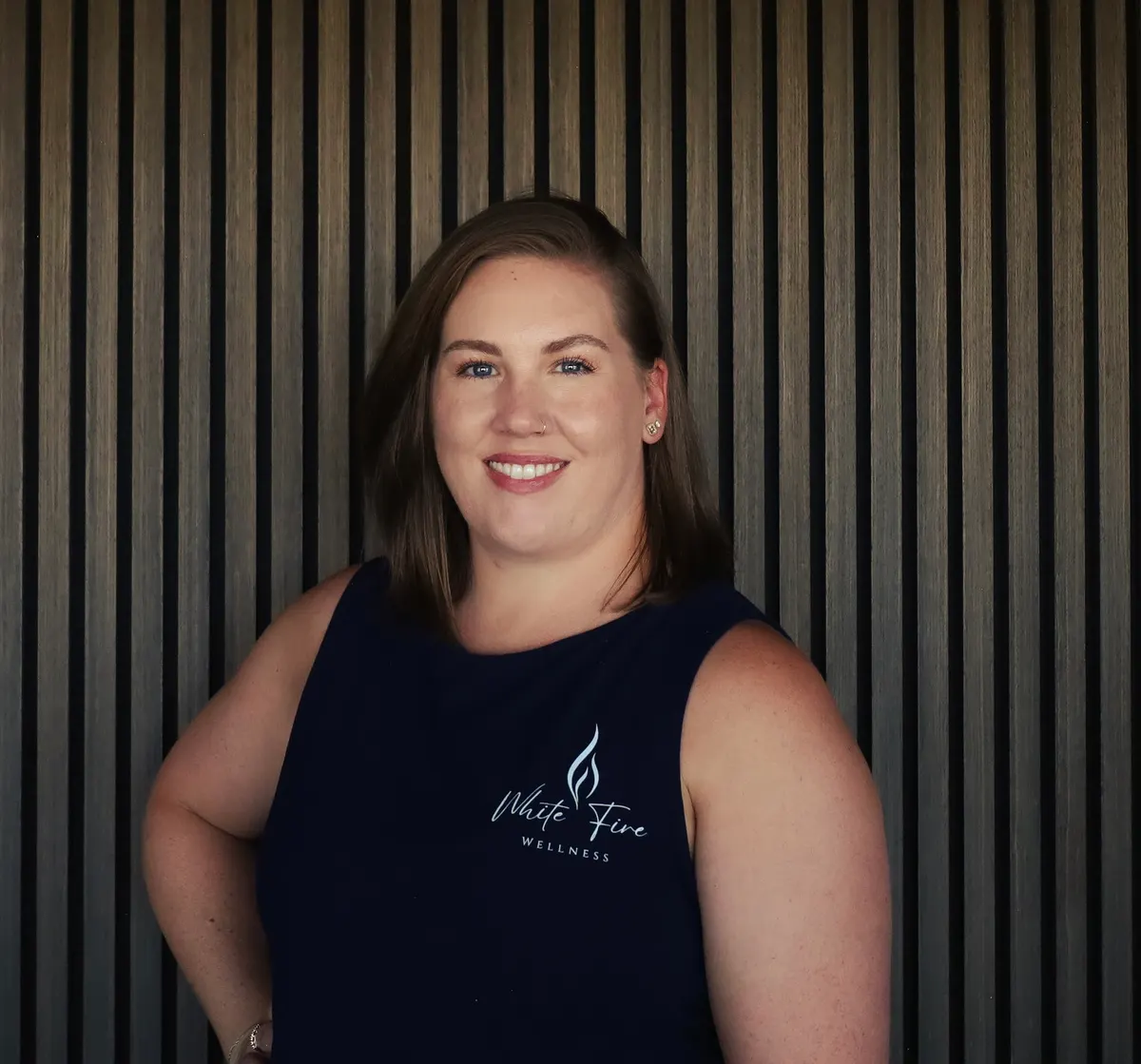 A woman with dark hair pulled back, in a navy White Fire tank top, smiles in front of a slatted wood wall.