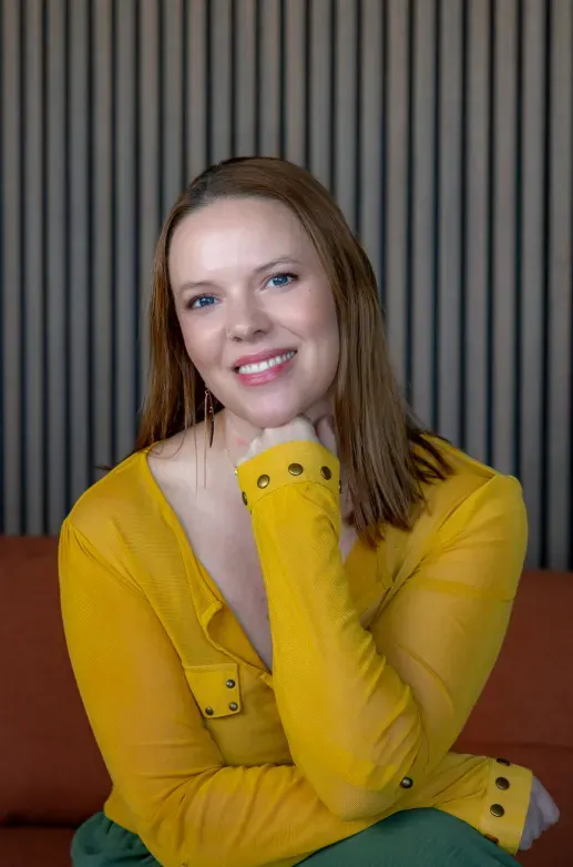 A woman with shoulder-length auburn hair and a mustard-yellow blouse smiles with her chin on her hand, in front of a slatted wood wall.
