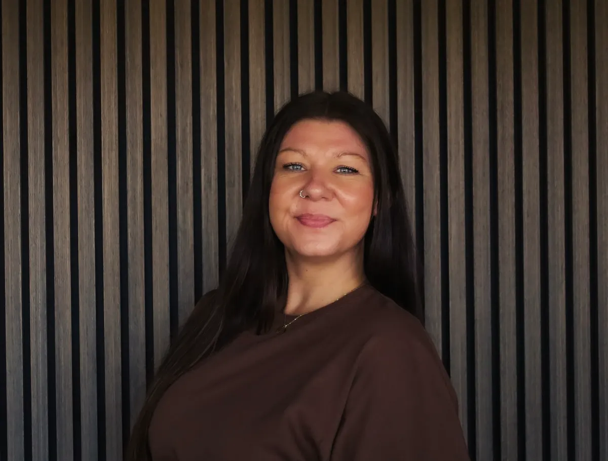 A woman with dark hair in a brown top smiles in front of a slatted wood wall.