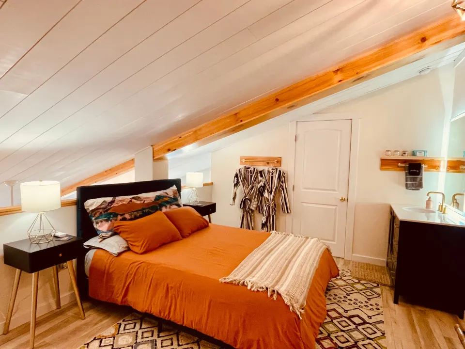 A bed with a rust-orange duvet under a sloped white plank ceiling and a pine beam, with two striped robes hanging beside a white door.