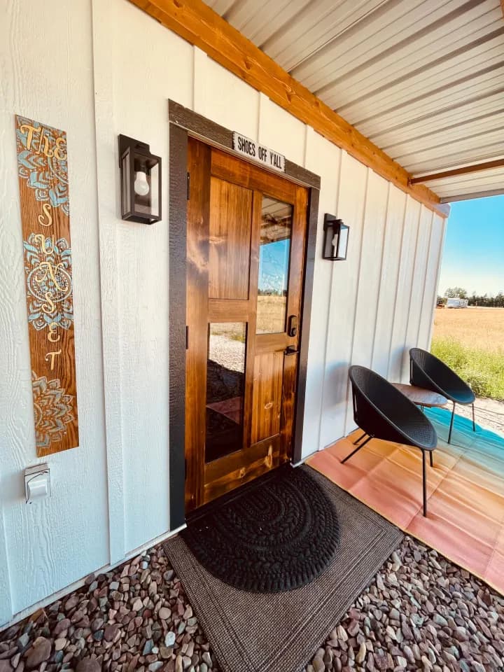 A wood-and-glass front door under a covered porch, with two black chairs on a pink and teal rug beside a summer field.