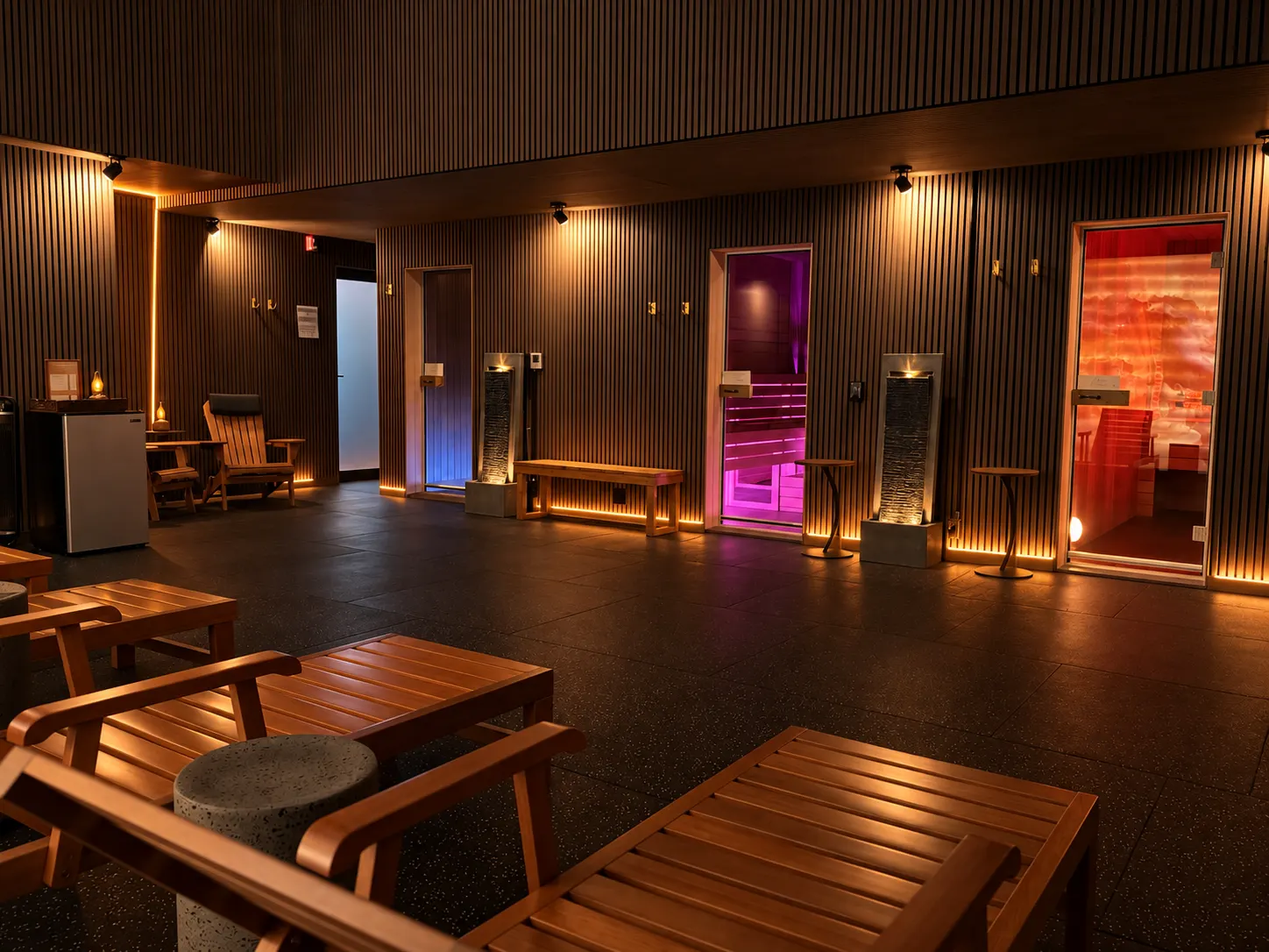 The thermal spa floor: wooden loungers and a low table on dark tile, slat-clad walls lit from beneath, and three sauna doors along the far wall glowing amber, magenta and red.