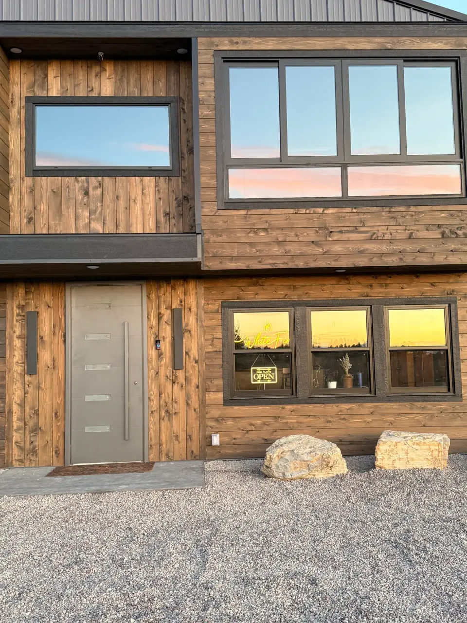 The front entrance at dusk, with wood siding, a tall gray door and a White Fire neon sign glowing in the window above two boulders.