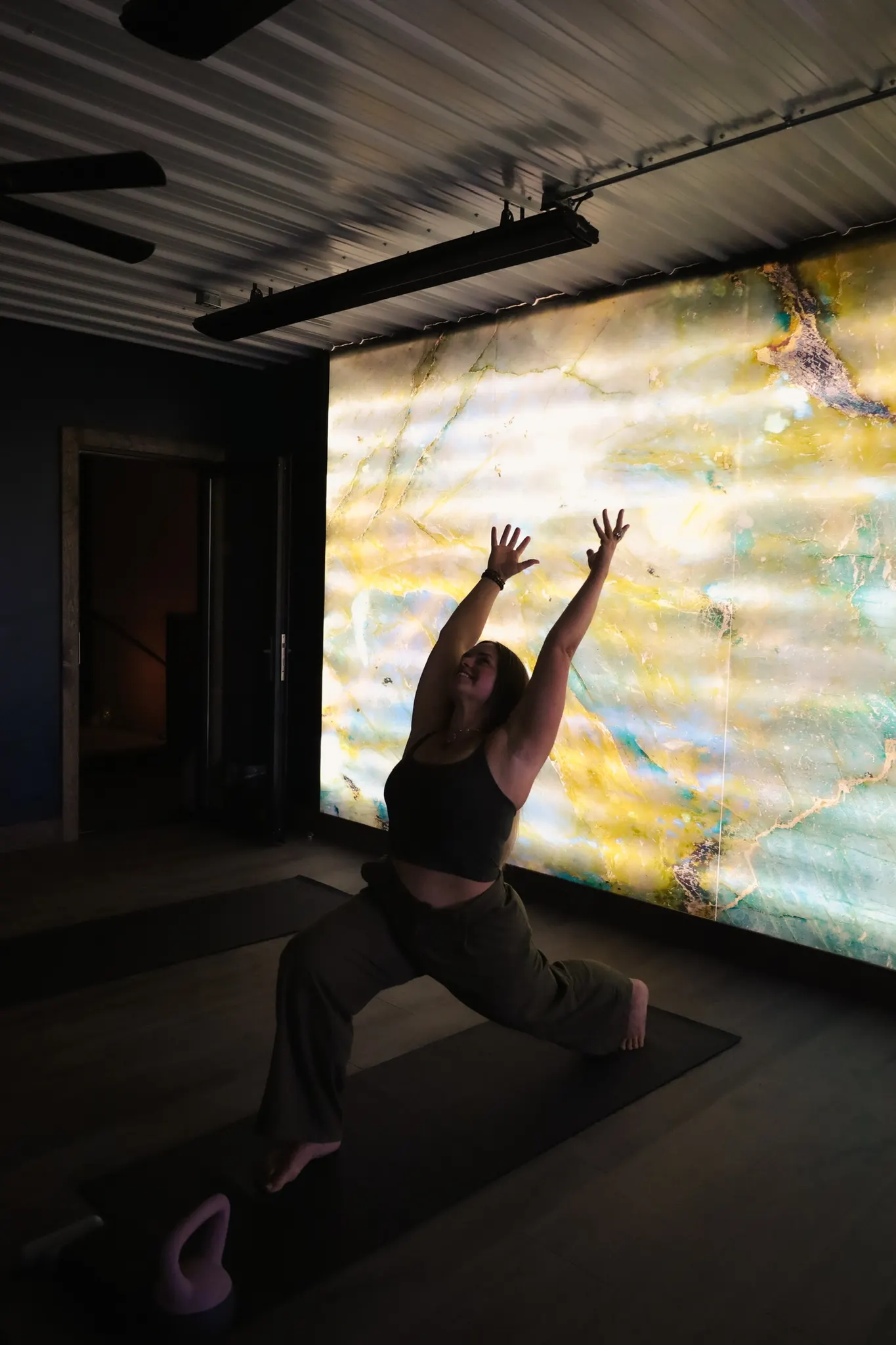 A woman in a high lunge with both arms raised, in silhouette against the glowing backlit stone wall of a dark studio.